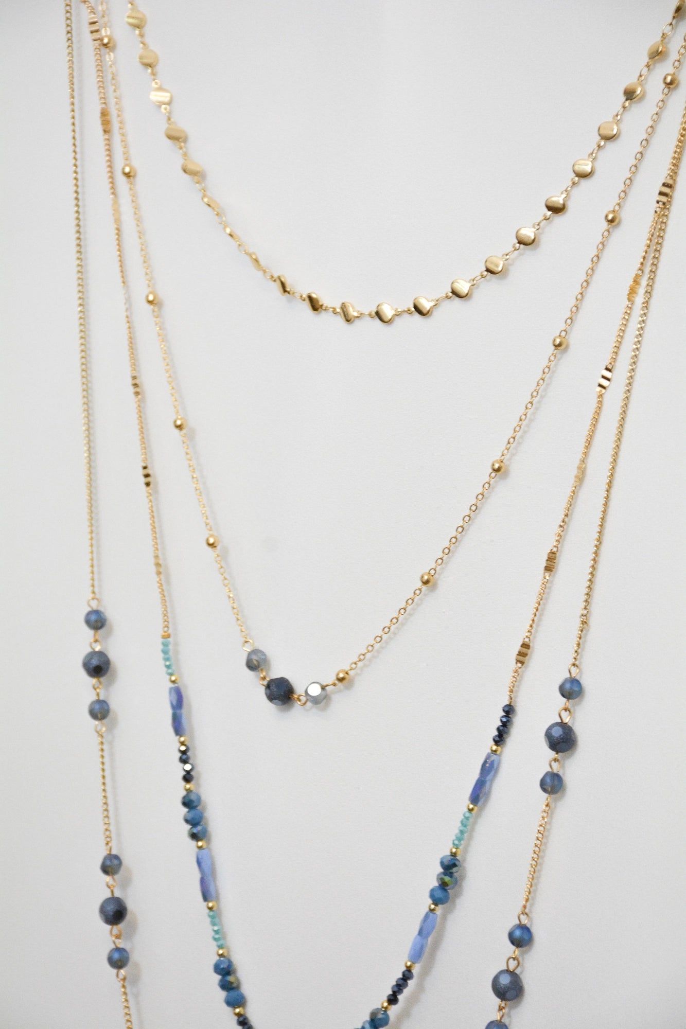 Detailaufnahme der Kette Mavi: erste Kette durchgehend goldfarben, zweite Kette mit blauen Glasperlen, Fokus auf die ersten beiden Ketten